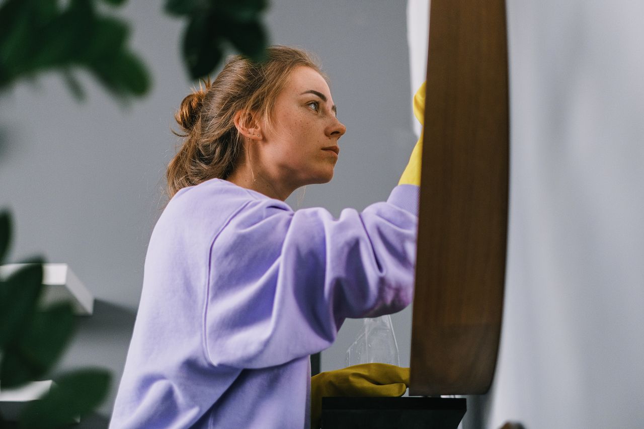 Side view of a young focused woman wearing latex gloves, standing and cleaning a mirror in a bright room during the day